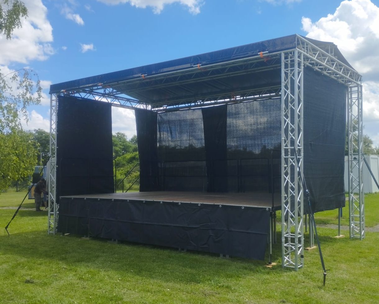 Bühne mit schwarzem Vorhang und blauem Dach in einer grünen Landschaft.