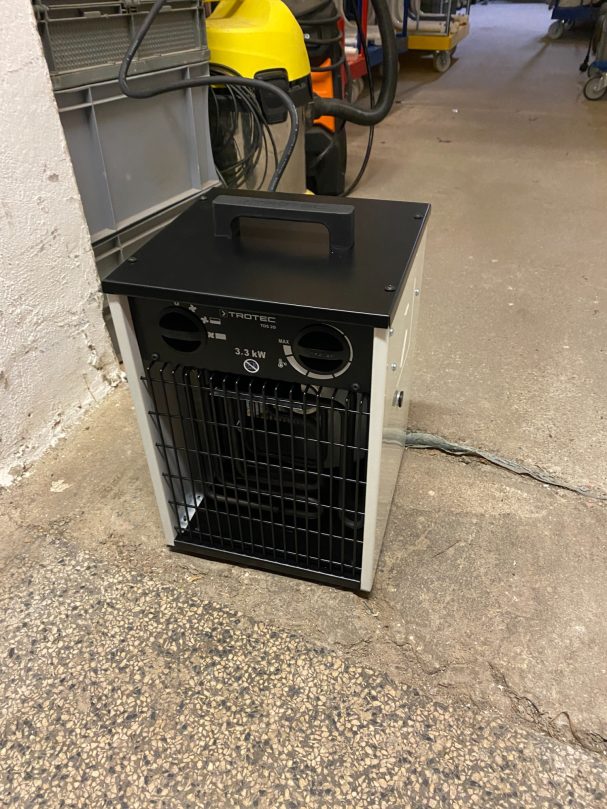 Schwarzer Heizlüfter mit Metallgehäuse und Bedienelementen, auf einem Betonboden.