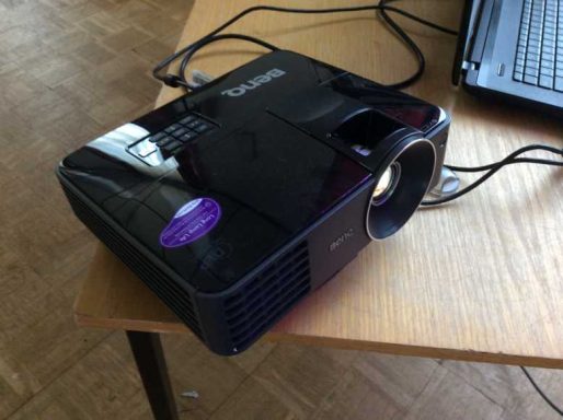 Ein schwarzer Projektor auf einem Tisch, angeschlossen mit Kabeln.