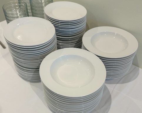 Teller mieten Hochgestapelte weiße Teller auf einem Tisch, im Hintergrund Glaswaren.